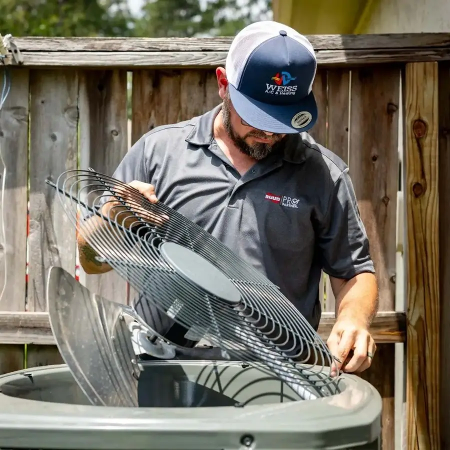 A Weiss Air Conditioning & Plumbing technician removing the top grille of an outdoor AC condenser to inspect and clean the fan blade.